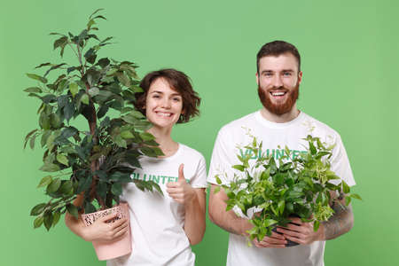 Smiling two young friends couple in white volunteer t-shirt isolated on green background. Voluntary free work assistance help charity grace teamwork concept. Hold green flowerpots, showing thumb up.の写真素材