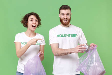 Excited friends couple in volunteer t-shirt isolated on green background. Voluntary free work assistance help charity grace teamwork concept. Picking up trash paper point index finger on garbage bags.の写真素材