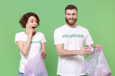 Shocked friends couple in volunteer t-shirt isolated on green background. Voluntary free work assistance help charity grace teamwork concept. Picking up trash paper in garbage bags put hand on cheek.の写真素材