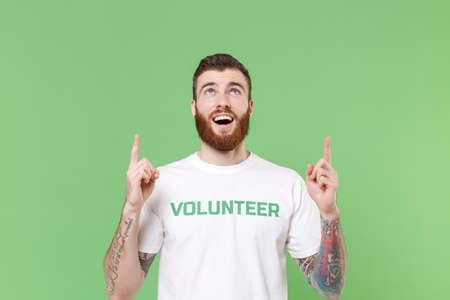 Excited young bearded man in white volunteer t-shirt isolated on pastel green background studio portrait. Voluntary free work assistance help charity grace teamwork concept. Pointing index fingers up.の写真素材