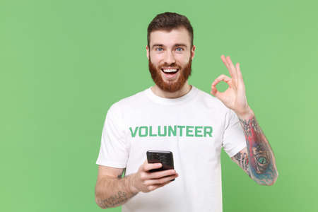 Cheerful young bearded man in white volunteer t-shirt isolated on pastel green background. Voluntary free work assistance help charity grace teamwork concept. Using mobile phone, showing OK gesture.の写真素材