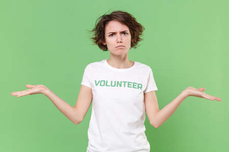 Displeased young brunette woman in white volunteer t-shirt isolated on pastel green wall background studio portrait. Voluntary free work assistance help charity grace teamwork concept. Spreading handsの写真素材