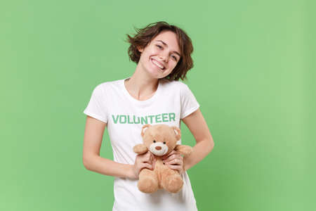 Pretty young brunette woman in white volunteer t-shirt isolated on pastel green background in studio. Voluntary free work assistance help charity grace teamwork concept. Holding teddy bear plush toy.の写真素材