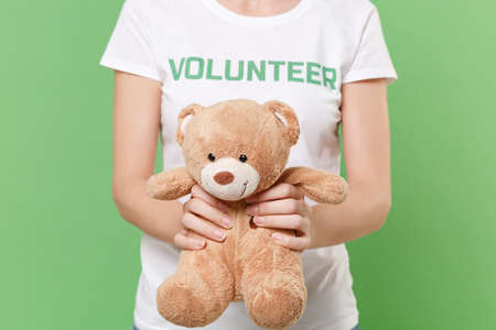 Cropped image of young woman in white volunteer t-shirt isolated on pastel green background in studio. Voluntary free work assistance help charity grace teamwork concept. Hold teddy bear plush toy.の写真素材