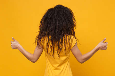 Back rear view of young african american woman girl in casual t-shirt posing isolated on yellow orange wall background studio portrait. People lifestyle concept. Mock up copy space. Showing thumbs up.の写真素材