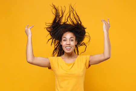 Funny young african american woman girl in casual t-shirt posing isolated on yellow orange background. People lifestyle concept. Mock up copy space. Have fun, throwing fluttering hair spreading hands.の写真素材