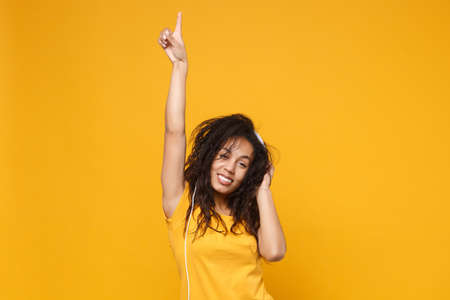 Smiling young african american woman in casual t-shirt posing isolated on yellow background. People lifestyle concept. Mock up copy space. Listen music with headphones, point index finger up, dancing.の写真素材