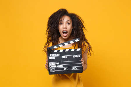 Shocked young african american woman girl in casual t-shirt posing isolated on yellow orange wall background. People lifestyle concept. Mock up copy space. Hold classic black film making clapperboard.の写真素材