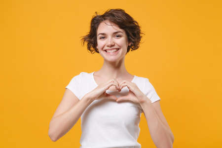 Smiling young brunette woman girl in white t-shirt posing isolated on yellow orange background in studio. People lifestyle concept. Mock up copy space. Showing shape heart with hands heart-shape sign.の写真素材