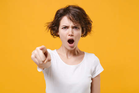 Angry young brunette woman girl in white t-shirt posing isolated on yellow orange background in studio. People lifestyle concept. Mock up copy space. Point index finger on camera swearing screaming.の写真素材