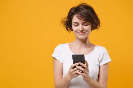 Smiling young brunette woman girl in white t-shirt posing isolated on yellow orange background studio portrait. People lifestyle concept. Mock up copy space. Using mobile phone, typing sms message.の写真素材