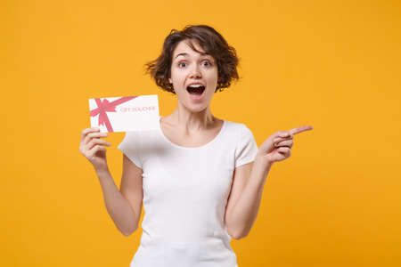 Shocked young brunette woman girl in white t-shirt posing isolated on yellow orange wall background. People lifestyle concept. Mock up copy space. Hold gift certificate pointing index finger aside.の写真素材