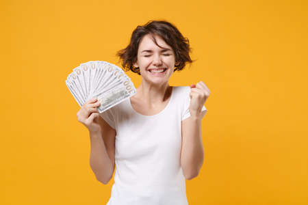 Joyful brunette woman in white t-shirt isolated on yellow background. People lifestyle concept. Mock up copy space. Hold fan of cash money in dollar banknotes doing winner gesture keeping eyes closed.の写真素材