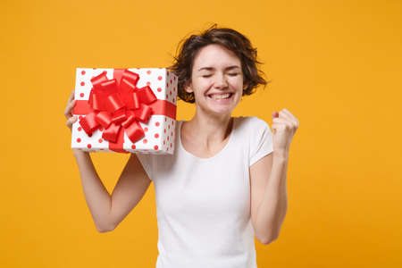 Happy woman in white t-shirt isolated on yellow background. Valentines Day Womens Day, birthday, holiday concept. Mock up copy space. Hold red present box with gift ribbon bow, doing winner gesture.の写真素材