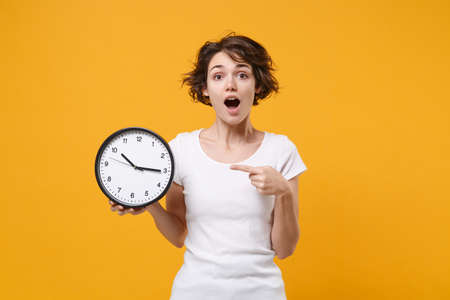 Shocked young brunette woman girl in white t-shirt posing isolated on yellow orange background studio portrait. People emotions lifestyle concept. Mock up copy space. Pointing index finger on clock.の写真素材