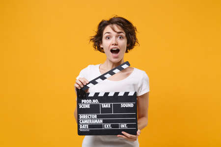 Shocked young brunette woman girl in white t-shirt posing isolated on yellow orange background in studio. People lifestyle concept. Mock up copy space. Hold classic black film making clapperboard.の写真素材