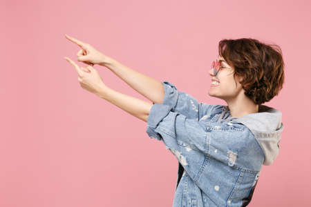 Side view of smiling young brunette woman girl in casual denim jacket eyeglasses posing isolated on pastel pink background. People lifestyle concept. Mock up copy space. Point index fingers aside up.の写真素材