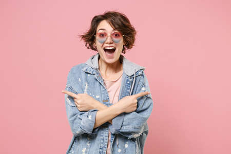 Surprised young brunette woman girl in casual denim jacket eyeglasses isolated on pastel pink background. People lifestyle concept. Mock up copy space. Pointing index fingers aside hold hands crossed.の写真素材