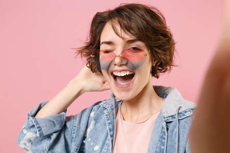 Close up of blinking young brunette woman in casual denim jacket eyeglasses posing isolated on...