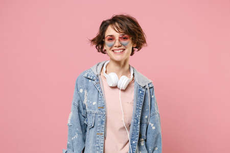 Smiling young brunette woman girl in casual denim jacket, eyeglasses with headphones posing isolated on pastel pink background in studio. People lifestyle concept. Mock up copy space. Looking camera.の写真素材