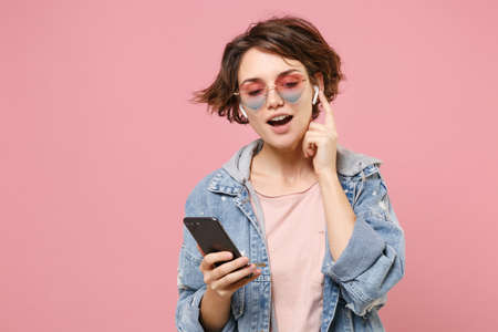 Pretty young brunette woman in casual denim jacket, eyeglasses posing isolated on pastel pink background. People lifestyle concept. Mock up copy space. Listen music with air pods, hold mobile phone.の写真素材