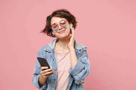 Charming young brunette woman in casual denim jacket, eyeglasses posing isolated on pastel pink background. People lifestyle concept. Mock up copy space. Listen music with air pods, hold mobile phone.の写真素材