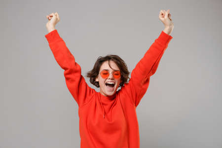 Funny young brunette woman girl in casual red hoodie, eyeglasses posing isolated on grey background in studio. People lifestyle concept. Mock up copy space. Doing winner gesture, keeping eyes closed.の写真素材