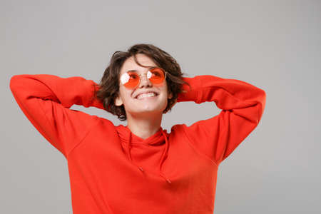 Pensive young brunette woman girl in casual red hoodie, eyeglasses posing isolated on grey background studio portrait. People lifestyle concept. Mock up copy space. Looking up with hands behind head.の写真素材