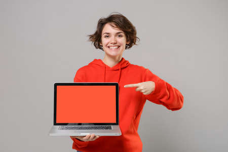 Smiling young brunette woman girl in casual red hoodie isolated on grey background. People lifestyle concept. Mock up copy space. Pointing index finger on laptop pc computer with blank empty screen.の写真素材