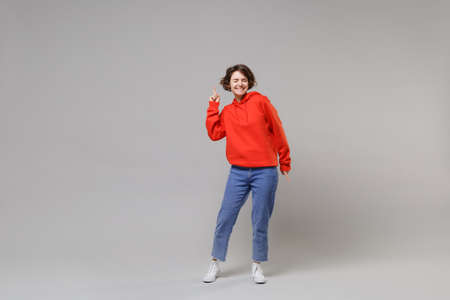 Smiling young brunette woman girl in casual red hoodie blue jeans posing isolated on grey wall background. People lifestyle concept. Mock up copy space. Pointing index finger up, keeping eyes closed.の写真素材