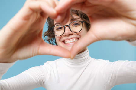 Smiling young brunette woman girl in casual white clothes, eyeglasses isolated on pastel blue background. People lifestyle concept. Mock up copy space. Showing shape heart with hands heart-shape sign.の写真素材