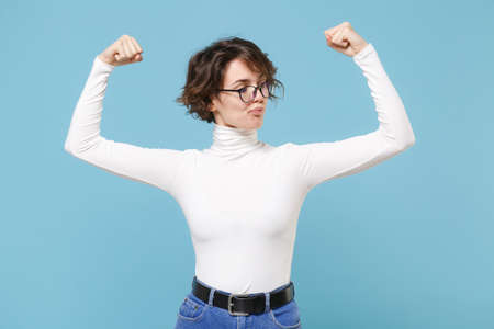Strong young brunette woman girl in casual white clothes, eyeglasses posing isolated on pastel blue background studio portrait. People lifestyle concept. Mock up copy space. Showing biceps, muscles.の写真素材