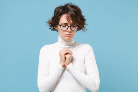Young brunette woman in casual white clothes eyeglasses posing isolated on pastel blue background in studio. People lifestyle concept. Mock up copy space. Holding hands folded in prayer, looking down.の写真素材