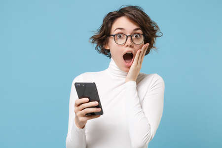 Shocked young brunette woman girl in casual white clothes, eyeglasses isolated on pastel blue background in studio. People lifestyle concept. Mock up copy space. Using mobile phone, put hand on cheek.の写真素材