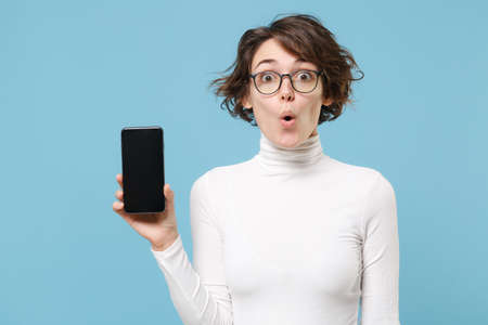 Shocked young brunette woman girl in casual white clothes eyeglasses posing isolated on pastel blue background. People lifestyle concept. Mock up copy space. Hold mobile phone with blank empty screen.の写真素材