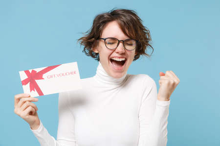 Happy young brunette woman girl in casual white clothes eyeglasses posing isolated on pastel blue background. People lifestyle concept. Mock up copy space. Hold gift certificate, doing winner gesture.の写真素材