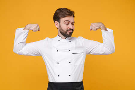 Strong young bearded male chef cook or baker man in white uniform shirt posing isolated on yellow...