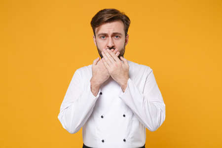 Concerned young bearded male chef cook or baker man in white uniform shirt posing isolated on yellow background studio portrait. Cooking food concept. Mock up copy space. Covering mouth with hands.の写真素材