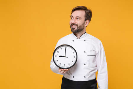 Smiling young bearded male chef cook or baker man in white uniform shirt posing isolated on yellow wall background studio portrait. Cooking food concept. Mock up copy space. Hold clock, looking aside.の写真素材