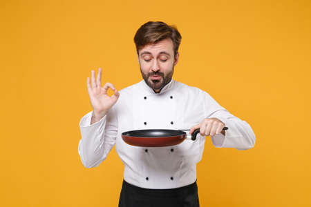 Young bearded male chef cook or baker man in white uniform shirt posing isolated on yellow wall background in studio. Cooking food concept. Mock up copy space. Holding frying pan, showing OK gesture.の写真素材