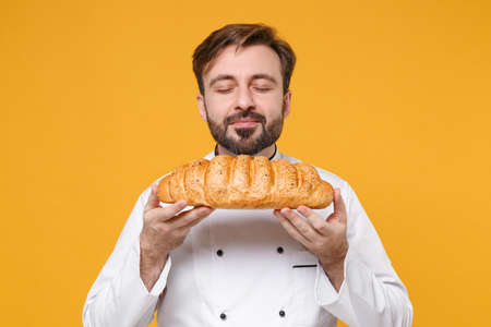 Young bearded male chef cook or baker man in white uniform shirt posing isolated on yellow background studio portrait. Cooking food concept. Mock up copy space. Sniffing bread, feeling food smell.の写真素材