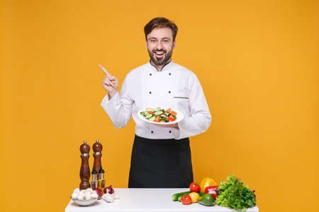 Excited bearded male chef cook or baker man in white uniform isolated on yellow background. Cooking food concept. Mock up copy space. Stand near table hold plate with salad pointing finger aside up.の写真素材