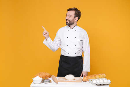 Smiling young bearded male chef cook or baker man in white uniform shirt isolated on yellow background. Cooking food concept. Mock up copy space. Cooking dough at table pointing index finger aside up.の写真素材
