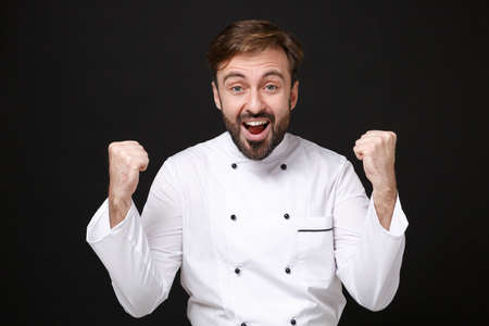 Surprised young bearded male chef cook or baker man in white uniform shirt posing isolated on black background studio portrait. Cooking food concept. Mock up copy space. Clenching fists like winner.の写真素材