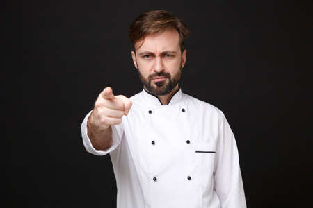 Dissatisfied young bearded male chef cook or baker man in white uniform shirt posing isolated on black background in studio. Cooking food concept. Mock up copy space. Pointing index finger on camera.の写真素材