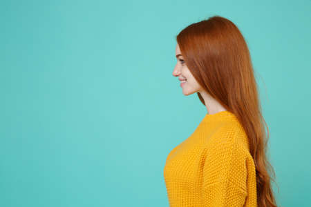 Side view of smiling young redhead woman girl in yellow sweater posing isolated on blue turquoise wall background studio portrait. People emotions lifestyle concept. Mock up copy space. Looking aside.の写真素材