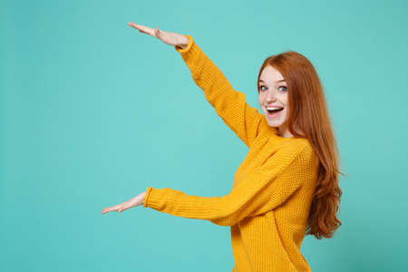 Excited young redhead woman girl in yellow sweater posing isolated on blue turquoise background. People lifestyle concept. Mock up copy space. Gesturing demonstrating size with vertical workspace.の写真素材