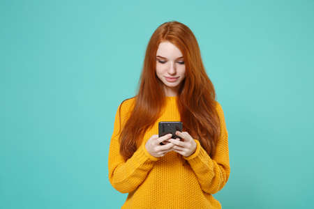 Pretty young redhead woman girl in yellow sweater posing isolated on blue turquoise background studio portrait. People lifestyle concept. Mock up copy space. Using mobile phone, typing sms message.の写真素材
