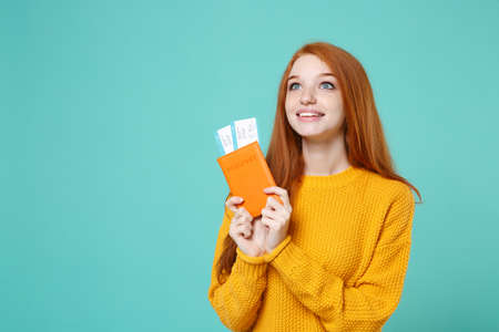 Excited young redhead girl in yellow sweater posing isolated on blue turquoise background in studio. People lifestyle concept. Mock up copy space. Hold passport boarding pass ticket looking aside up.の写真素材