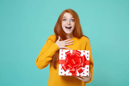 Surprised redhead girl in yellow sweater posing isolated on blue turquoise background. Valentines Day Womens Day, birthday concept. Hold white red present box with gift ribbon bow put hand on chest.の写真素材